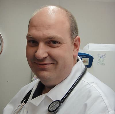 A man in a white lab coat with a stethoscope around his neck stands indoors, smiling at the camera.