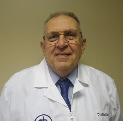 Older man wearing glasses, a white lab coat, blue shirt, and tie, standing in front of a plain beige wall, smiling at the camera.