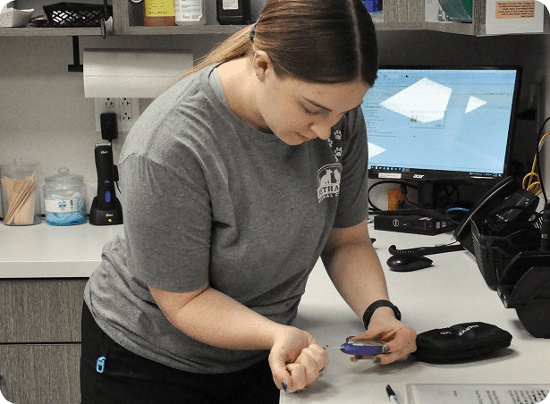 A woman in a gray T-shirt checks her blood sugar with a glucometer at a desk with a computer and medical supplies.