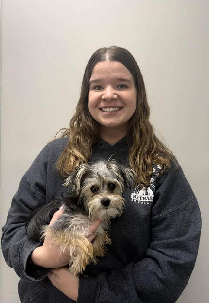 Woman with long brown hair wearing a dark sweatshirt holds a small black and gray dog in front of a plain light-colored wall.