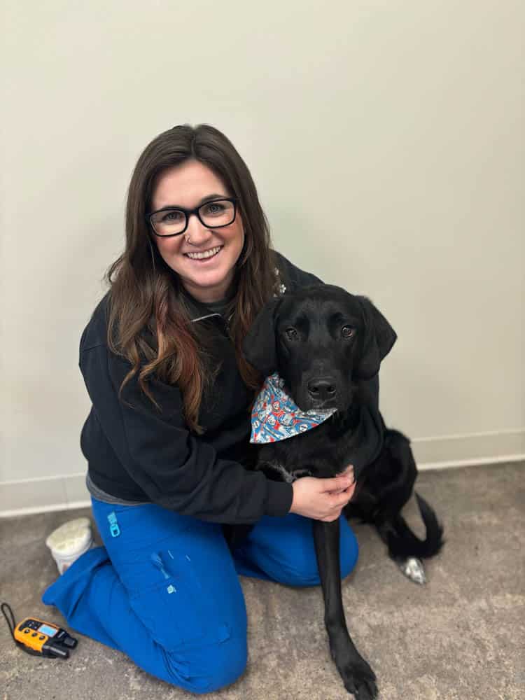 A woman in glasses and blue scrubs kneels next to a black dog wearing a colorful bandana. The woman is smiling, and they are indoors against a plain wall.