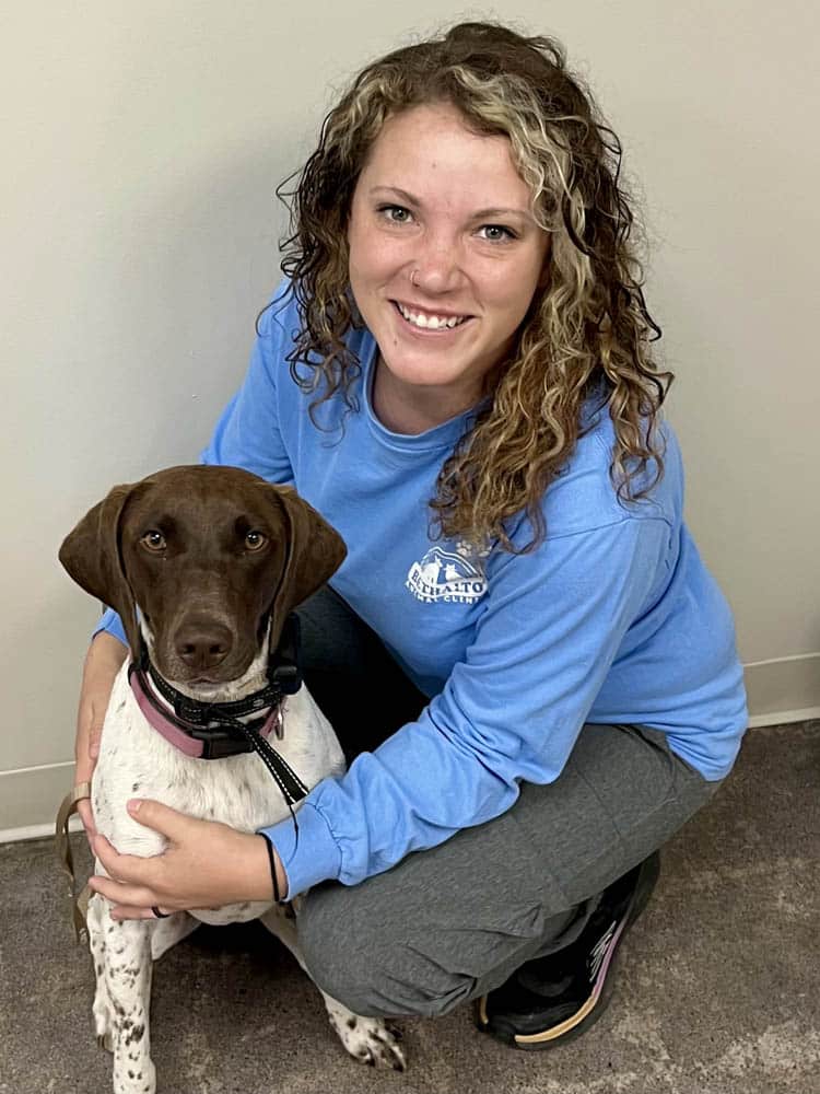 Woman with curly hair wearing a blue shirt crouches and smiles at the camera while holding a brown and white dog sitting beside her indoors on a carpeted floor.