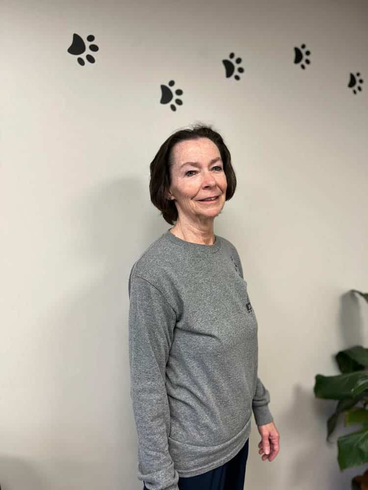 A woman in a gray sweatshirt stands indoors near a wall decorated with black paw print decals and a green plant in the corner.