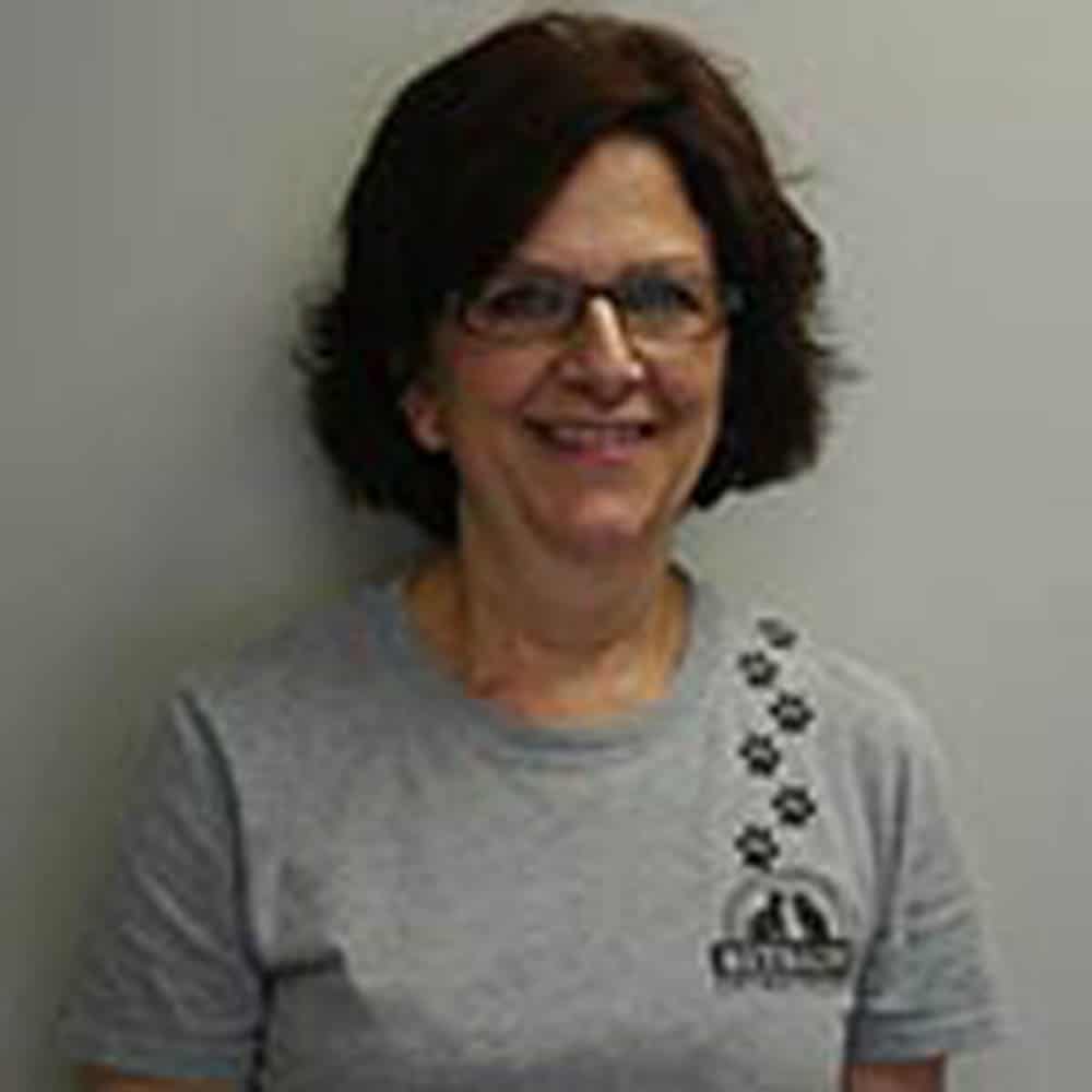 A woman with short brown hair, wearing glasses and a gray T-shirt with a logo and paw prints, stands against a plain background.